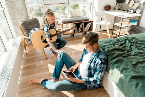 high angle view of man using digital tablet while girlfriend playing acoustic guitar at home - Fotoğraf, Görsel