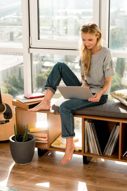 smiling young woman using laptop while sitting at window at home - Photo, Image