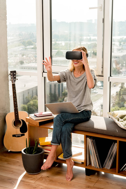 young woman in vr headset with laptop sitting at window at home - Fotó, kép
