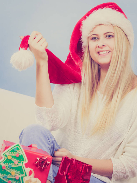 Christmas time concept. Happy teen blonde girl wearing santa claus hat preparing gifts for xmas, sitting at home on couch, cozy holiday interior - Zdjęcie, obraz