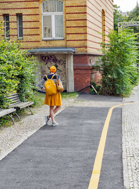 BERLIN, GERMANY - JULY 13, 2018: Urban scene with young woman with yellow hairs, backpack dressed in yellow skirt stands in front of yellow house and road marking line in Mitte district. - Photo, Image