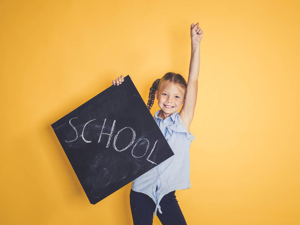 cute schoolgirl smiling and looking at camera while holding blackboard with word school and posing in front of orange background  - Фото, изображение