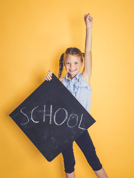 blond schoolgirl holding blackboard with word school while standing with raised hand on orange background  - 写真・画像