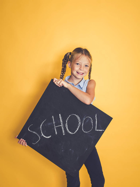 cute schoolgirl smiling and looking at camera while holding blackboard with word school and posing in front of orange background  - Foto, Imagen