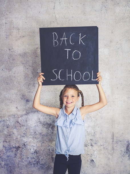 schoolgirl holding blackboard with words back to school in front of concrete background - Photo, Image