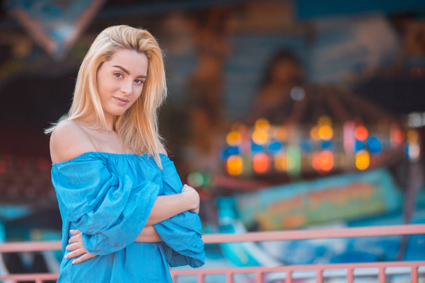 Portrait of happy young woman with blond hair in bright blue top against the backdrop of a carousel. Summer portrait of a girl on a bright background - Valokuva, kuva