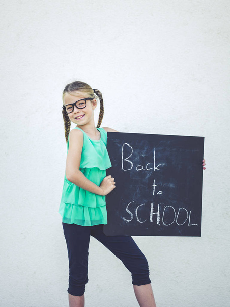 όμορφο κορίτσι με τα γυαλιά με πλεξούδες κρατώντας μαυροπίνακα με λόγια πίσω στο σχολείο  - Φωτογραφία, εικόνα