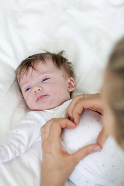 Młoda matka trzyma się za ręce w kształcie serca podczas masowania słodkie małe dziecko na łóżku - Zdjęcie, obraz