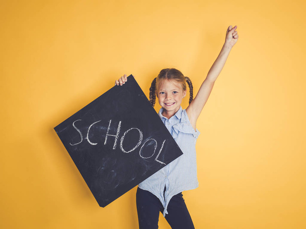blond schoolgirl holding blackboard with word school while standing with raised hand on orange background  - Φωτογραφία, εικόνα