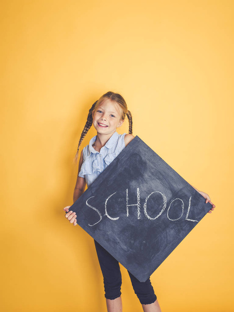 blond schoolgirl looking out from behind chalkboard with text school and posing in front of orange background  - 写真・画像