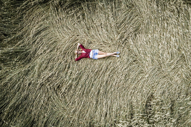 carefree beautiful woman wearing checkered shirt and denim short lying in rye field at sunny day, top view  - Photo, image