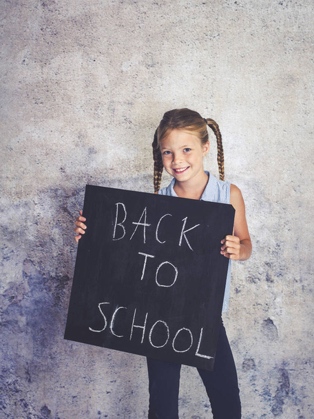 colegiala sosteniendo pizarra con palabras de vuelta a la escuela en frente de fondo de hormigón
 - Foto, imagen