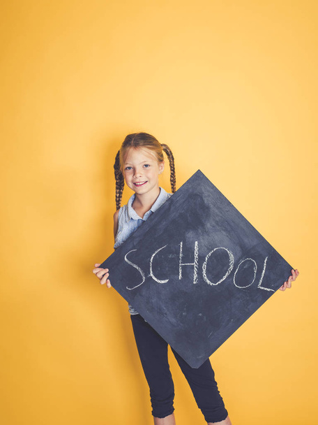 blond schoolgirl looking out from behind chalkboard with text school and posing in front of orange background  - Photo, Image