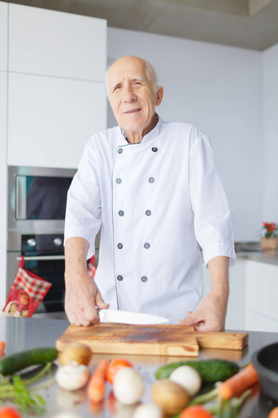Portrait of a smiling aged man with knife in the kitchen who preparing for dinner. Grandad with grey hair is slicing vegetables. - Photo, image
