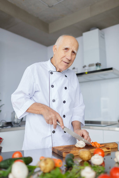 A smiling aged man with a knife in the light kitchen. Grandad with grey hair is slicing vegetables for a dinner. - Valokuva, kuva