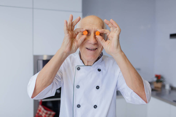 The elderly chef man with gray hair puts two pieces of carrot to the eyes in the kitchen. Useful vegetables - Fotografie, Obrázek