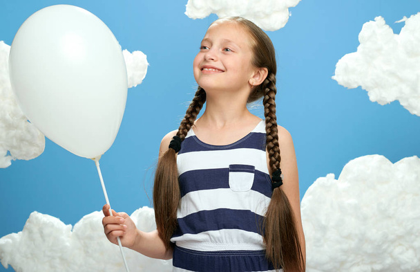 Mädchen in gestreiftem Kleid posiert vor blauem Hintergrund mit Baumwollwolken, weißem Luftballon, dem Konzept von Sommer und Glück - Foto, Bild