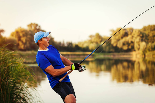 Jonge man visserij op rivier bij zonsondergang. Happy enthousiast fiserman trekken staaf met vis. Vangen van proces - Foto, afbeelding