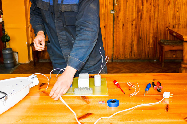 Un electricista trabajador trabaja, recoge el circuito eléctrico de una gran lámpara de calle blanca con cables, un relé en una fábrica de plantas industriales
 - Foto, Imagen