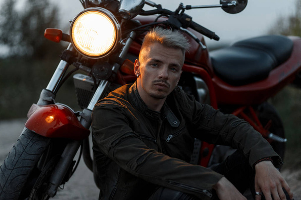 handsome hipster man with a hairstyle in a fashionable military khaki jacket sits near a motorcycle in the nature - Foto, Imagem