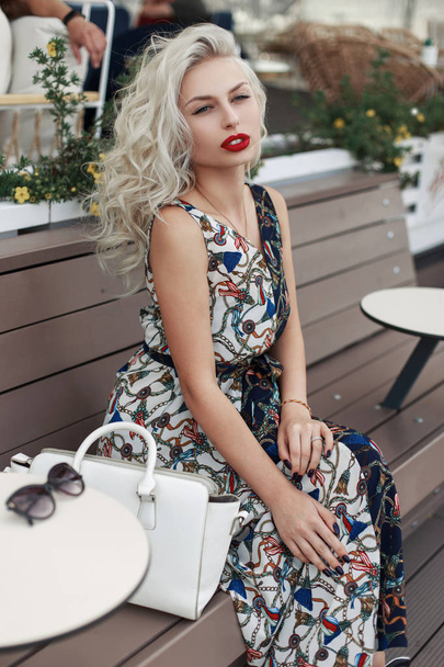 Mujer modelo joven bastante caliente de moda con un peinado en un vestido elegante con un patrón con una bolsa y gafas de sol sentado en un banco de madera en la ciudad
 - Foto, Imagen