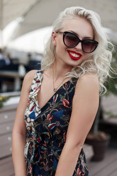 Αστεία όμορφη νεαρή κοπέλα με ένα χαμόγελο στα γυαλιά ηλίου σε ένα μοντέρνο φόρεμα με μοτίβο στο δρόμο - Φωτογραφία, εικόνα