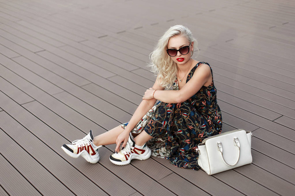 Cool stylish woman with sunglasses in a fashionable dress with patterns and shoes with a bag sits on the wooden floor of the deck - Photo, Image