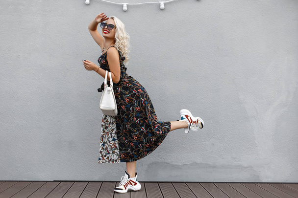 красивая забавная молодая стильная девушка с модной белой сумкой в винтажном платье позирует возле серой стены
 - Фото, изображение
