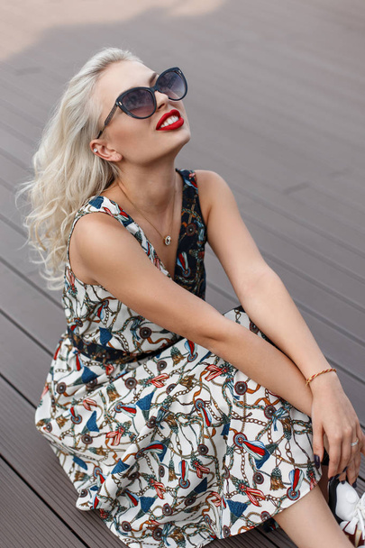 Κομψό hipster όμορφη νεαρή γυναίκα με γυαλιά ηλίου σε ένα vintage φόρεμα κάθεται σε μια ξύλινη προβλήτα στην παραλία - Φωτογραφία, εικόνα