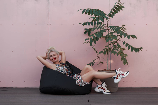 Chica joven feliz en vestido de moda se ríe y descansa en una bolsa de silla cerca de una pared rosa
 - Foto, Imagen