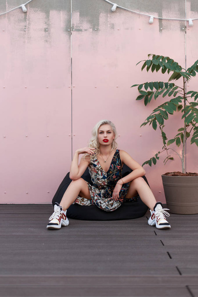 Bella donna alla moda modello giovane in un abito elegante con un modello in scarpe da ginnastica si siede in una borsa sedia vicino a una parete rosa
 - Foto, immagini