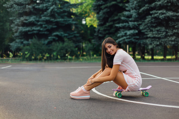 Portrait of a smiling charming brunette female sitting on her skateboard on a basketball court. Happy woman with trendy look taking break during sunset. - Photo, image