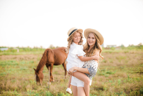 A beautiful young girl, with light curly hair in a straw hat with a little sister hugging and laughing near horses, in the countryside, warm autumn on a sunny day - Foto, immagini