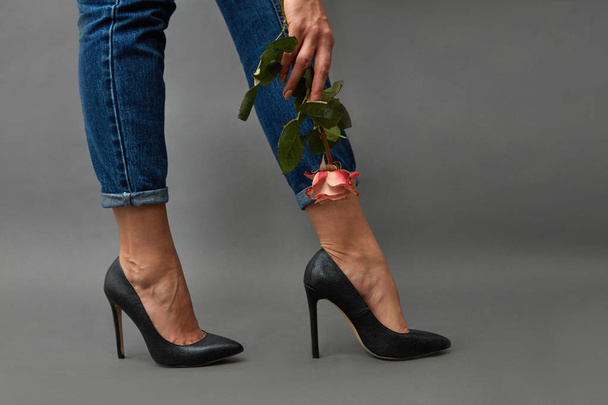 Elegant girl legs in jeans and high-heeled shoes, a girl's hand with a tattoo is holding a pink flower around a dark background with copy space. - Фото, зображення