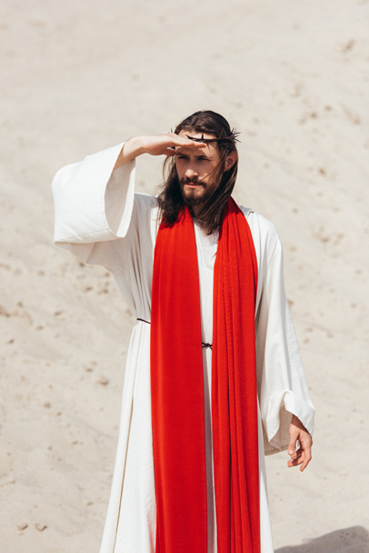 Jesus in robe, red sash and crown of thorns looking away in desert - Photo, Image