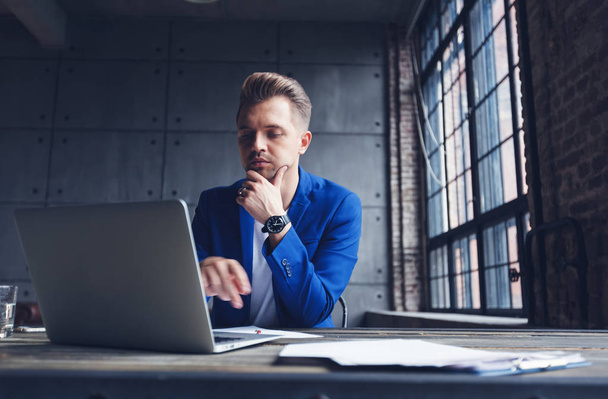 Young attractive business man in blue suit is sitting in the office at the desk with laptop and phone, marketing and communication management - Zdjęcie, obraz