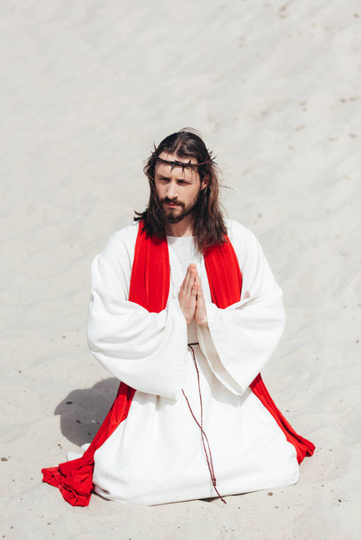 Jesus in robe, red sash and crown of thorns standing on knees and praying in desert, looking away - Photo, Image