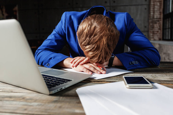 Jeune homme d'affaires en costume bleu, bouleversé, fatigué et déprimé assis au bureau avec ordinateur portable et téléphone, marketing et gestion de la communication
 - Photo, image