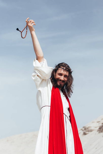 happy Jesus in robe, red sash and crown of thorns holding rosary in raised hand in desert - Fotó, kép