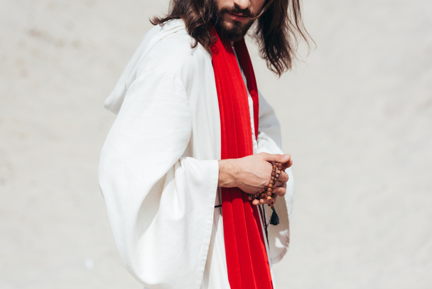 bijgesneden afbeelding Jezus in gewaad en rode sjerp holding rozenkrans in woestijn  - Foto, afbeelding