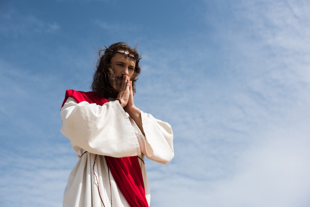 visão de baixo ângulo de Jesus em roupão e faixa vermelha orando contra o céu azul
 - Foto, Imagem