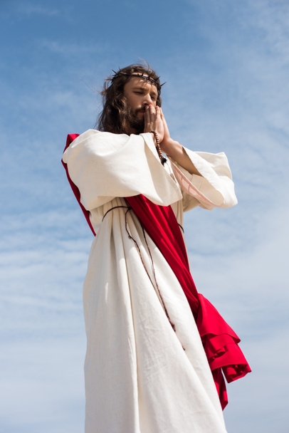 low angle view of Jesus in robe, red sash and crown of thorns holding rosary and praying against blue sky - Photo, Image