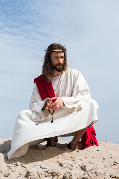 Jézus a ruhámat, a piros káva és a koronát a tövisek a rózsafüzért imádkozott, és guggolva a homokos sivatagban-hegy - Fotó, kép