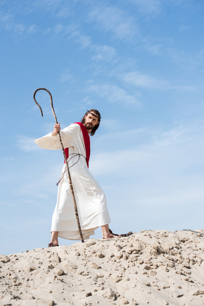 low angle view of Jesus in robe, red sash and crown of thorns walking on sandy hill with staff in desert - Photo, Image