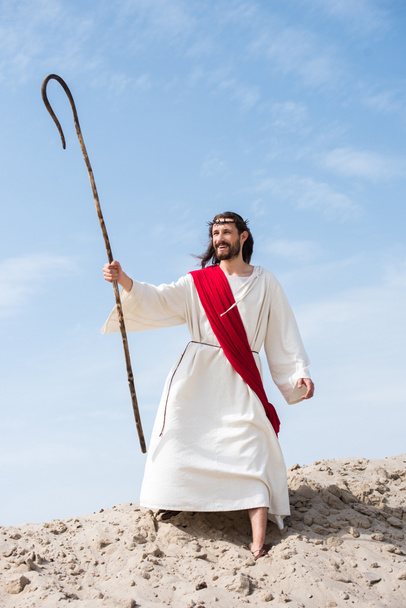 Jesus feliz em roupão, faixa vermelha e coroa de espinhos de pé com cajado de madeira no deserto e olhando para longe
 - Foto, Imagem