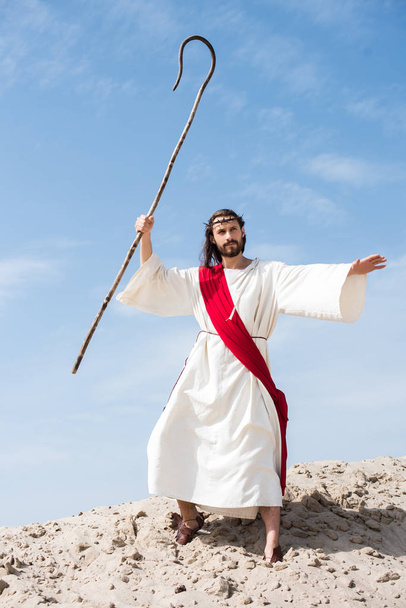 Jesus in Robe, roter Schärpe und Dornenkrone schwingen Holzstab in der Wüste - Foto, Bild
