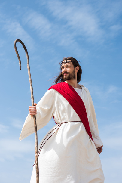 cheerful Jesus in robe, red sash and crown of thorns standing with wooden staff in desert - Foto, Imagen