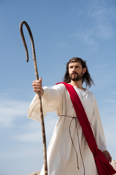 Jesus in robe, red sash and crown of thorns standing with wooden staff in desert - Photo, Image