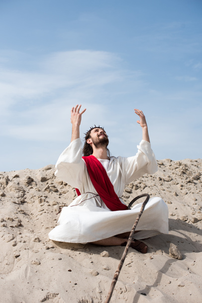 alegre Jesús en túnica y faja roja sentado en posición de loto con las manos levantadas y hablando con Dios en la arena en el desierto
 - Foto, imagen