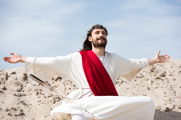 Jésus en robe, ceinture rouge et couronne d'épines assis en position lotus avec les bras ouverts et les yeux fermés dans le désert
 - Photo, image